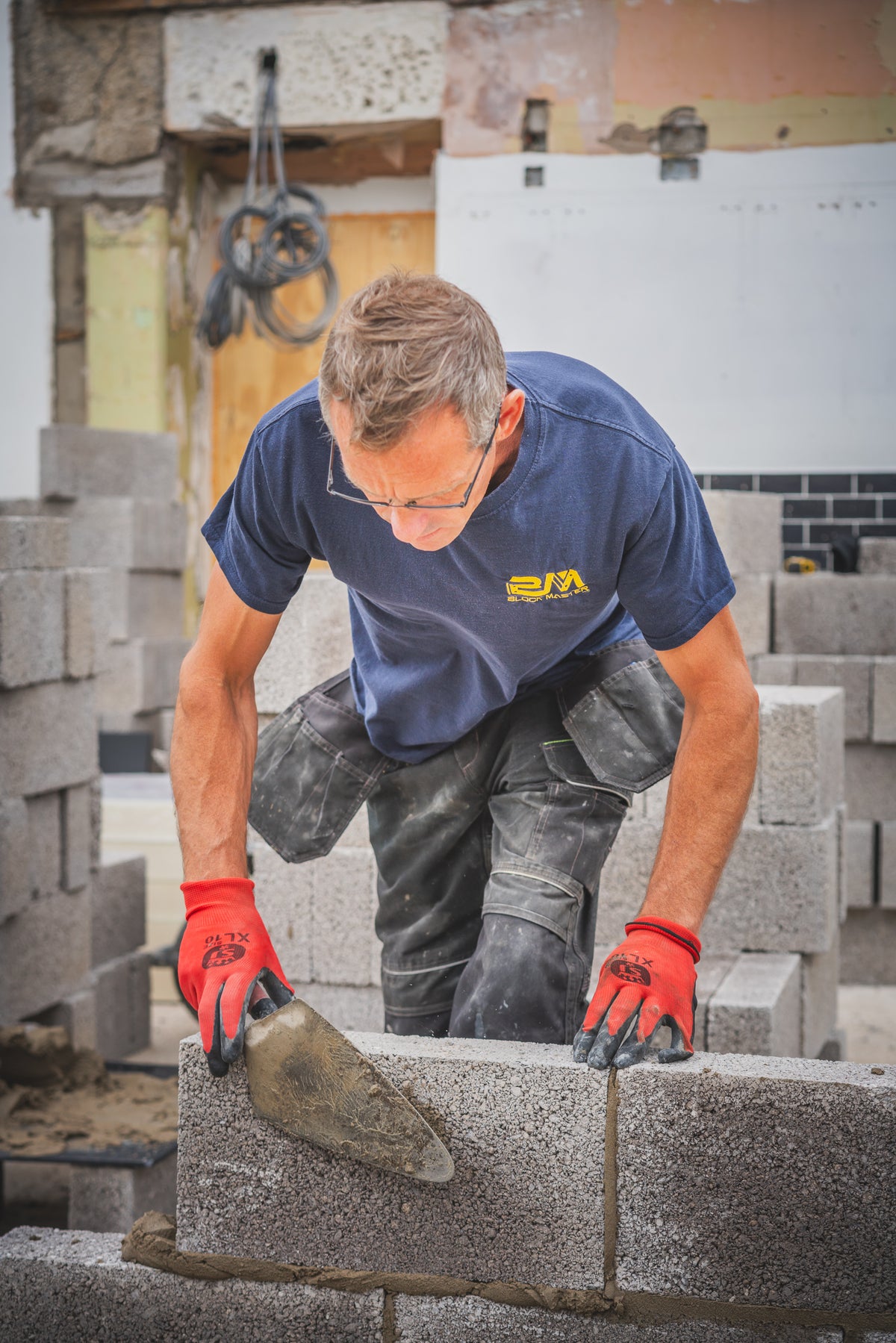 A man wearing a blue t-shirt with a yellow logo and red work gloves is building a wall with gray concrete blocks on a construction site.